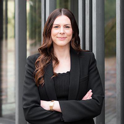 Porträt von Laura Hausmann: Frau im schwarzen Blazer mit verschränkten Armen vor Glasfassade, lächelt in die Kamera